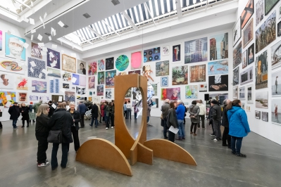 Eine Ausstellungshalle mit vielen Besucher*innen, die sich unterhalten und Kunstwerke betrachten. Die Wände sind dicht mit Gemä