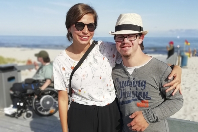 Junger Mensch mit Down-Syndrom, der gemeinsam mit seiner Reisebegleitung am Strand steht. Im Hintergrund ein Rollstuhlfahrer.