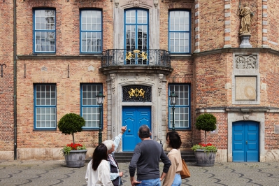 Das Bild zeigt eine Stadtführung am Rathaus. Die Gruppe steht vor einer großen Türe und blickt auf die Fassade.
