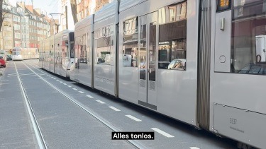 Straßenbahnen fahren eine Straße mit Hochhäusern entlang. Am unteren Bildrand erscheint die Texteinblendung 